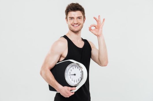 Image of happy young sportsman standing isolated over white background holding scales showing okay gesture. Looking at camera.